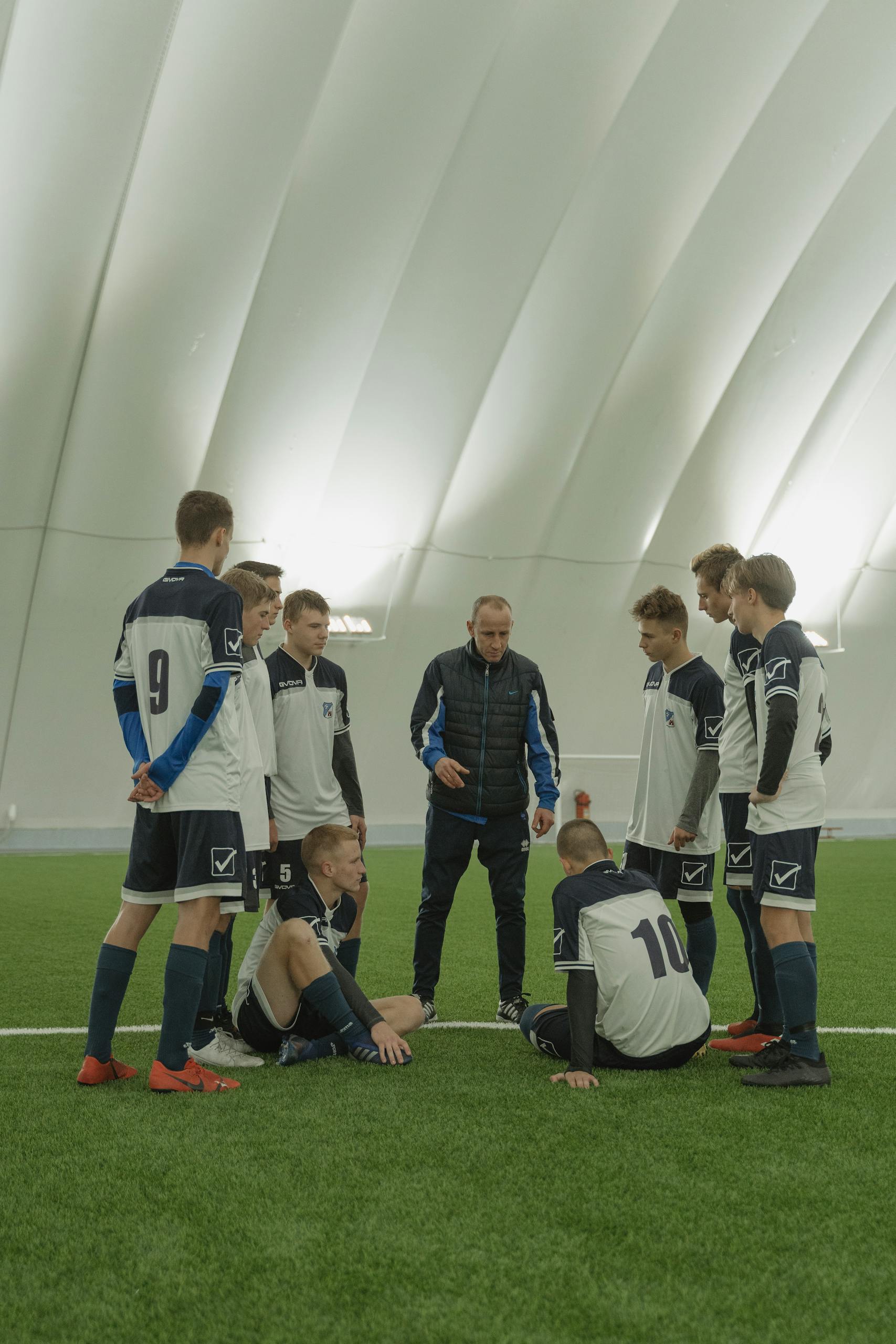 A coach giving a motivational talk to a youth soccer team inside an indoor stadium.