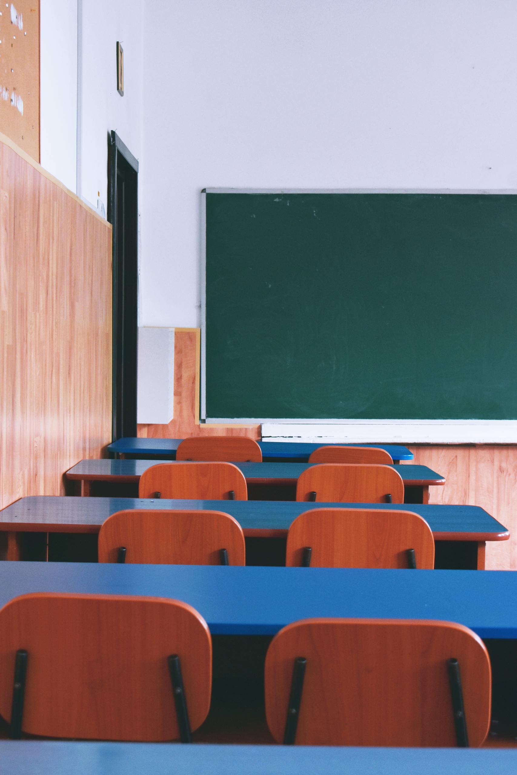 A serene view of an empty classroom with wooden desks, chairs, and a green chalkboard, capturing a quiet educational atmosphere.