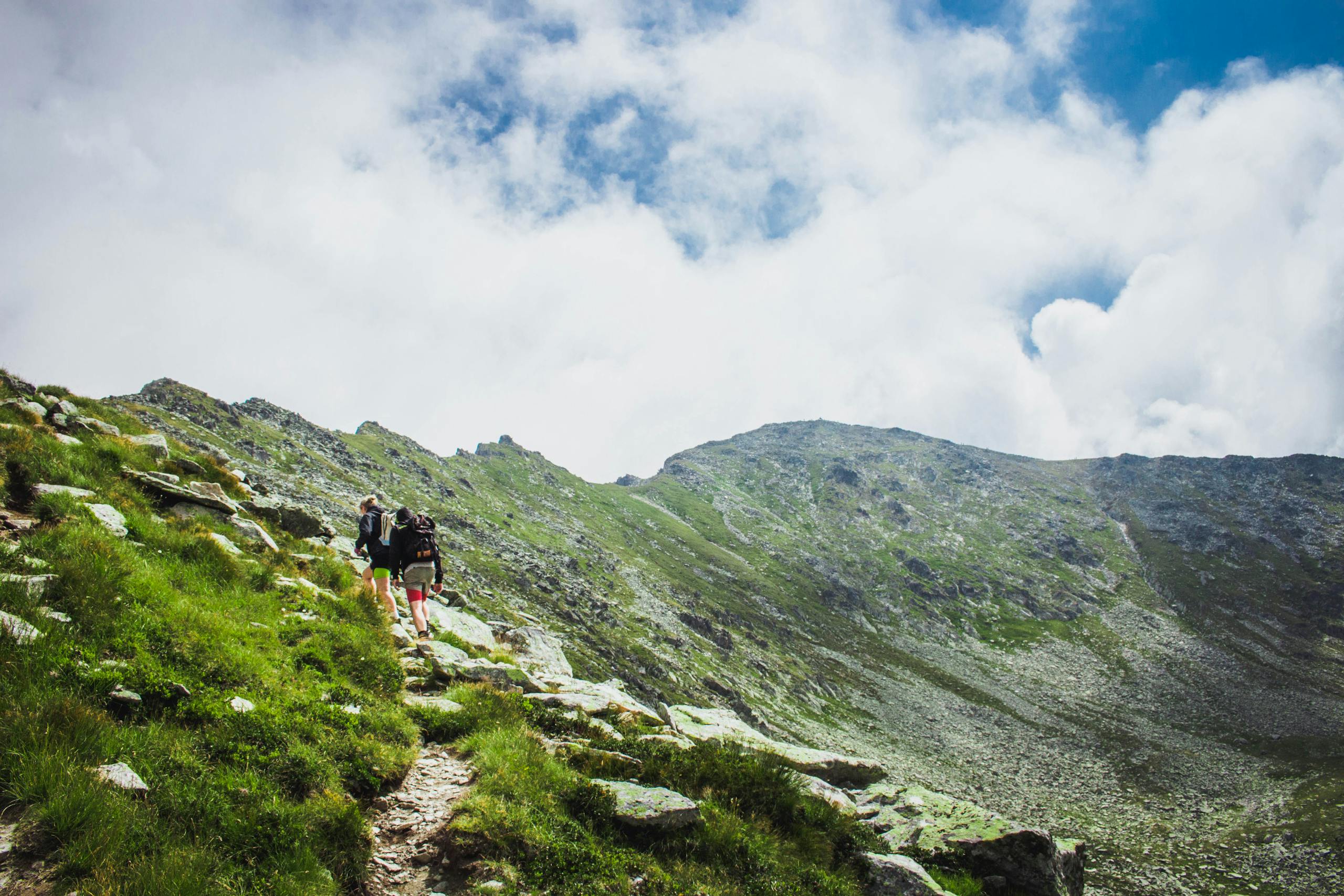 Two hikers explore lush green mountains under a bright sky in Romania, capturing the essence of adventure.