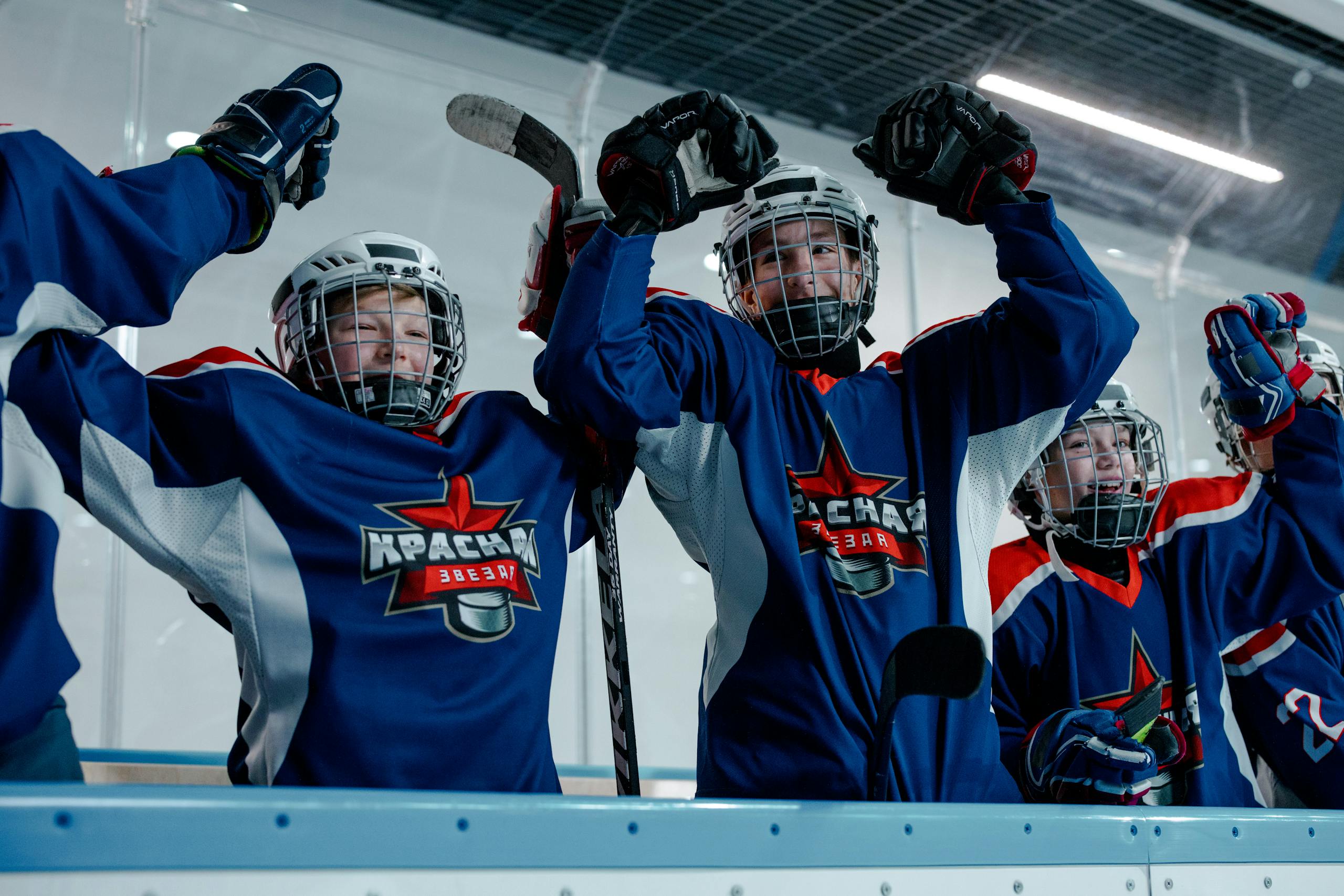Youth ice hockey team celebrates a triumphant moment indoors with raised hands and helmets.
