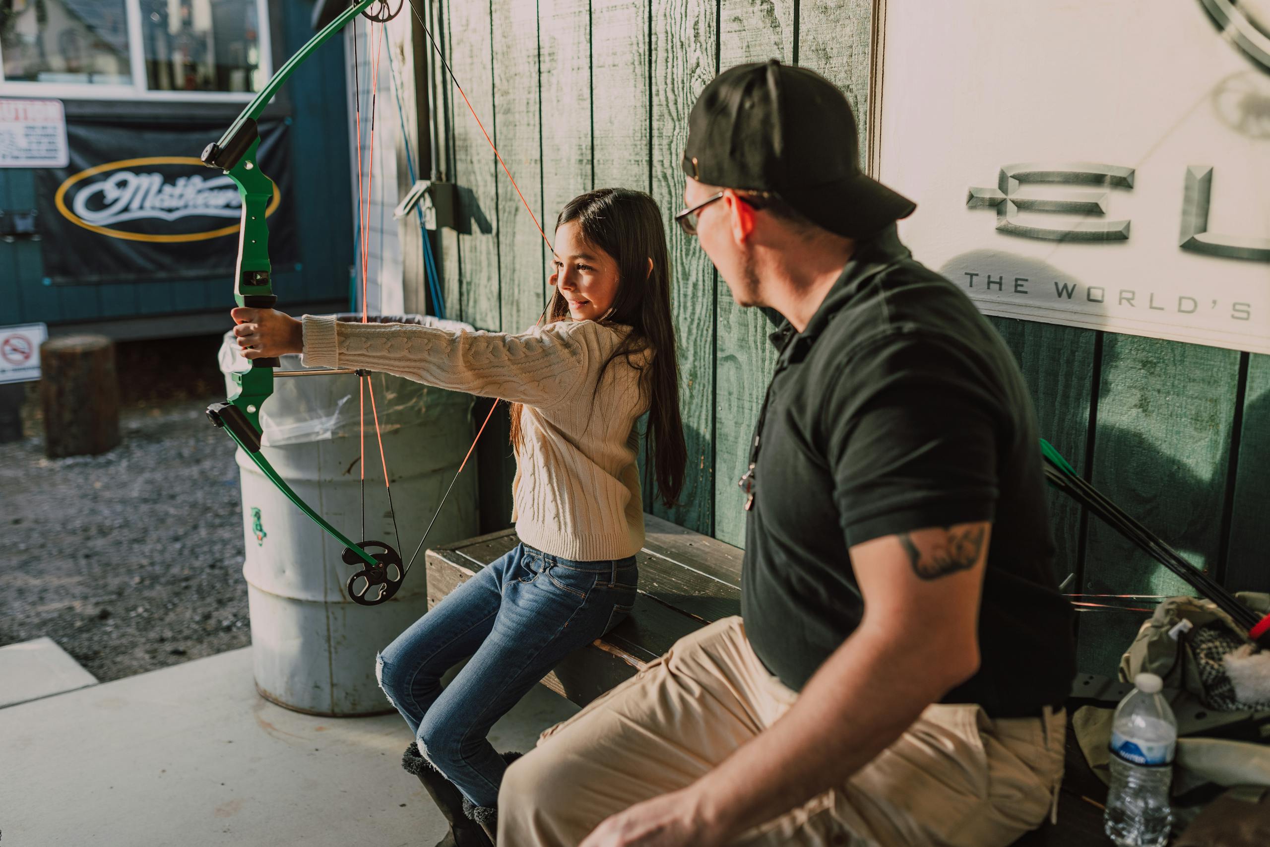 A young girl practices archery with a mentor outdoors, holding a compound bow.