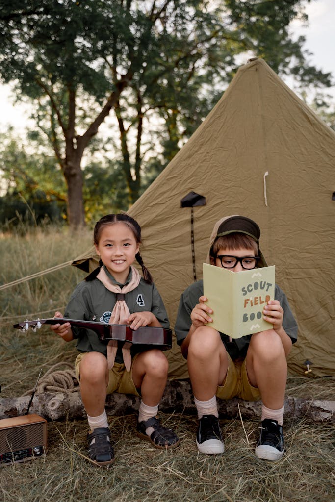 Children enjoying a scouting camp, reading and playing ukulele beside a tent.
