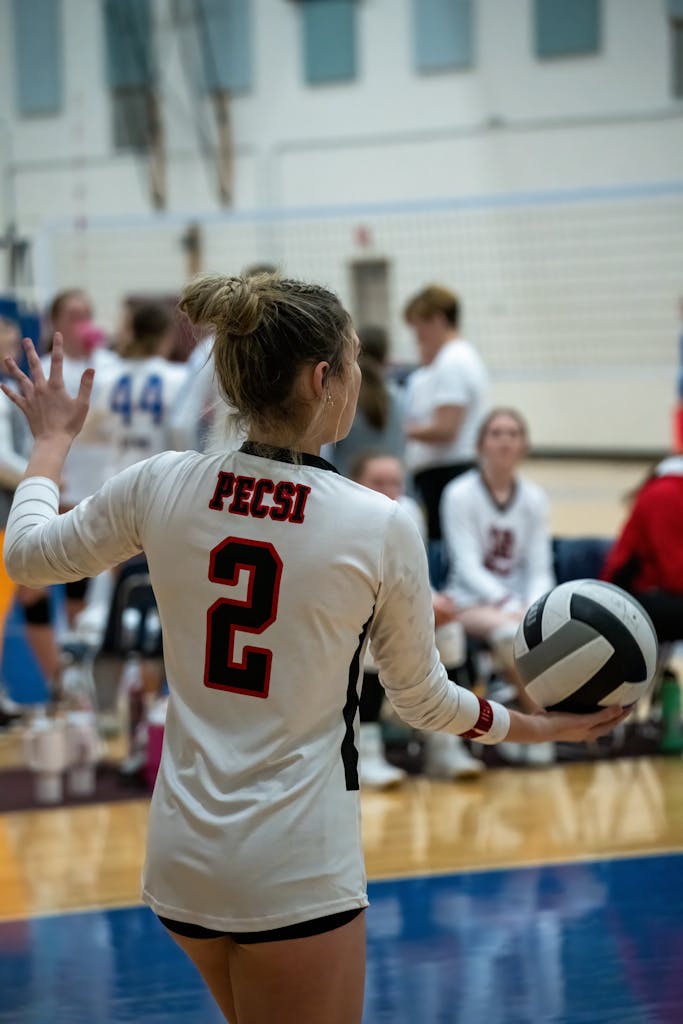 Teenage girl in volleyball match holding ball, preparing to serve. Indoor court setting.